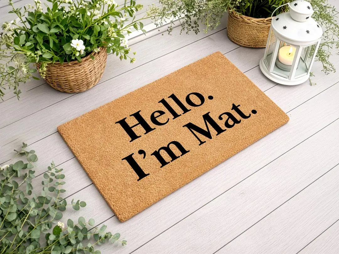 Coir doormat with a Hello I'm Mat message on a white wood deck with a lantern and flower baskets