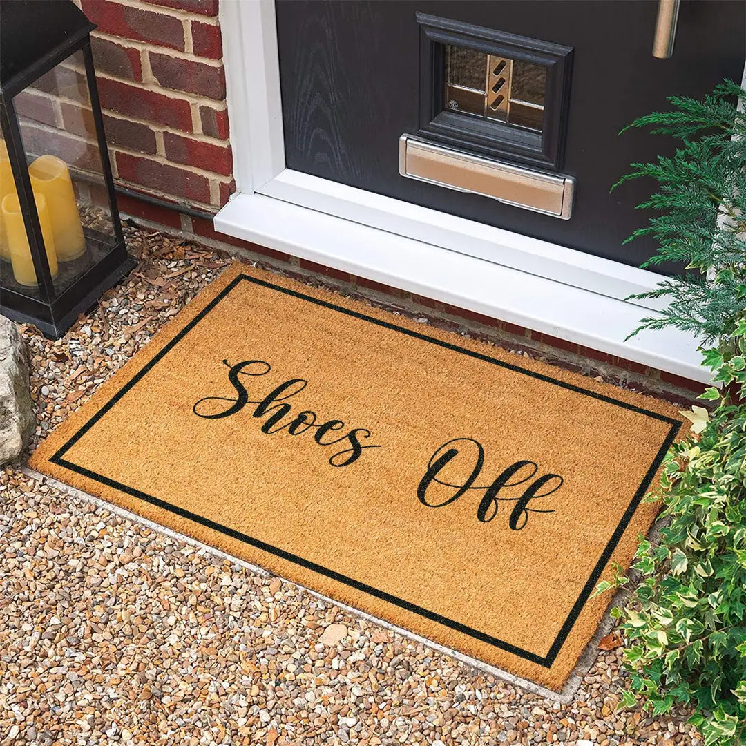 Rectangular coir doormat with Shoes Off script lettering and a black border at a dark front door on gravel