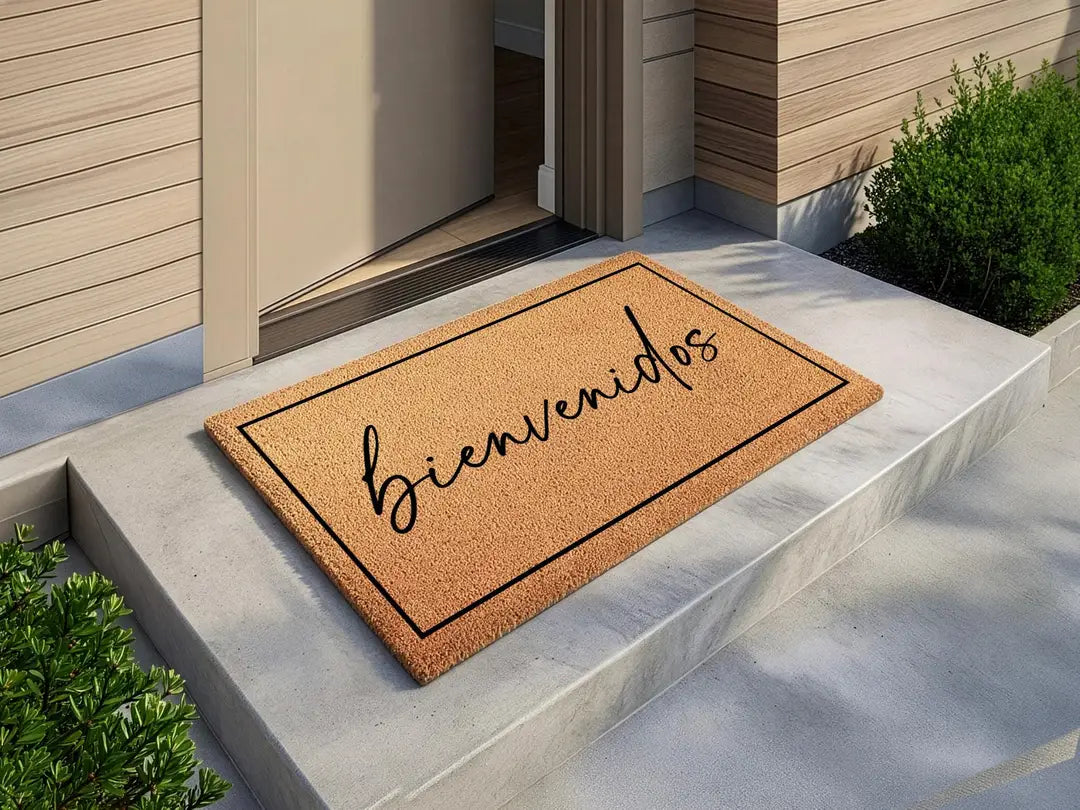 Coir doormat with a framed bienvenidos script message on a concrete step beside a door and shrubs