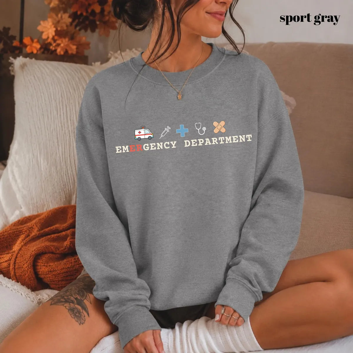 Sport Grey crewneck sweatshirt with printed an ambulance, syringe, cross, stethoscope and bandage above the words emergency department, worn by a model.