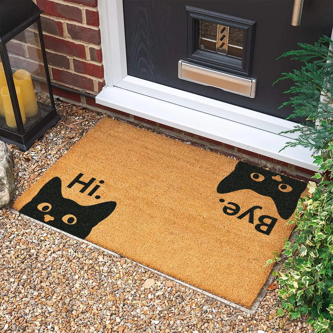 Rectangular coir doormat with Hi and Bye lettering and two peeking black cats on gravel in front of a black door