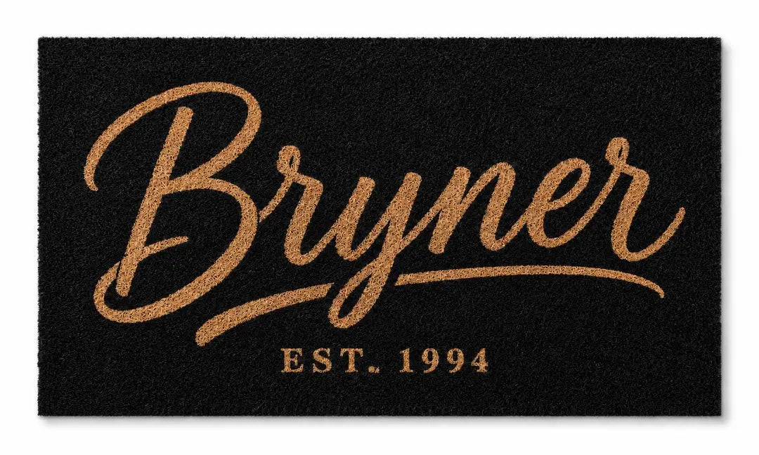 Flat view of a black faux coir doormat with a large gold-tone script surname and established year