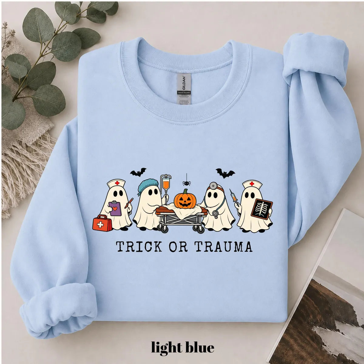Light Blue crewneck sweatshirt with printed nurse ghosts with a pumpkin and the words trick or trauma, flat lay view.