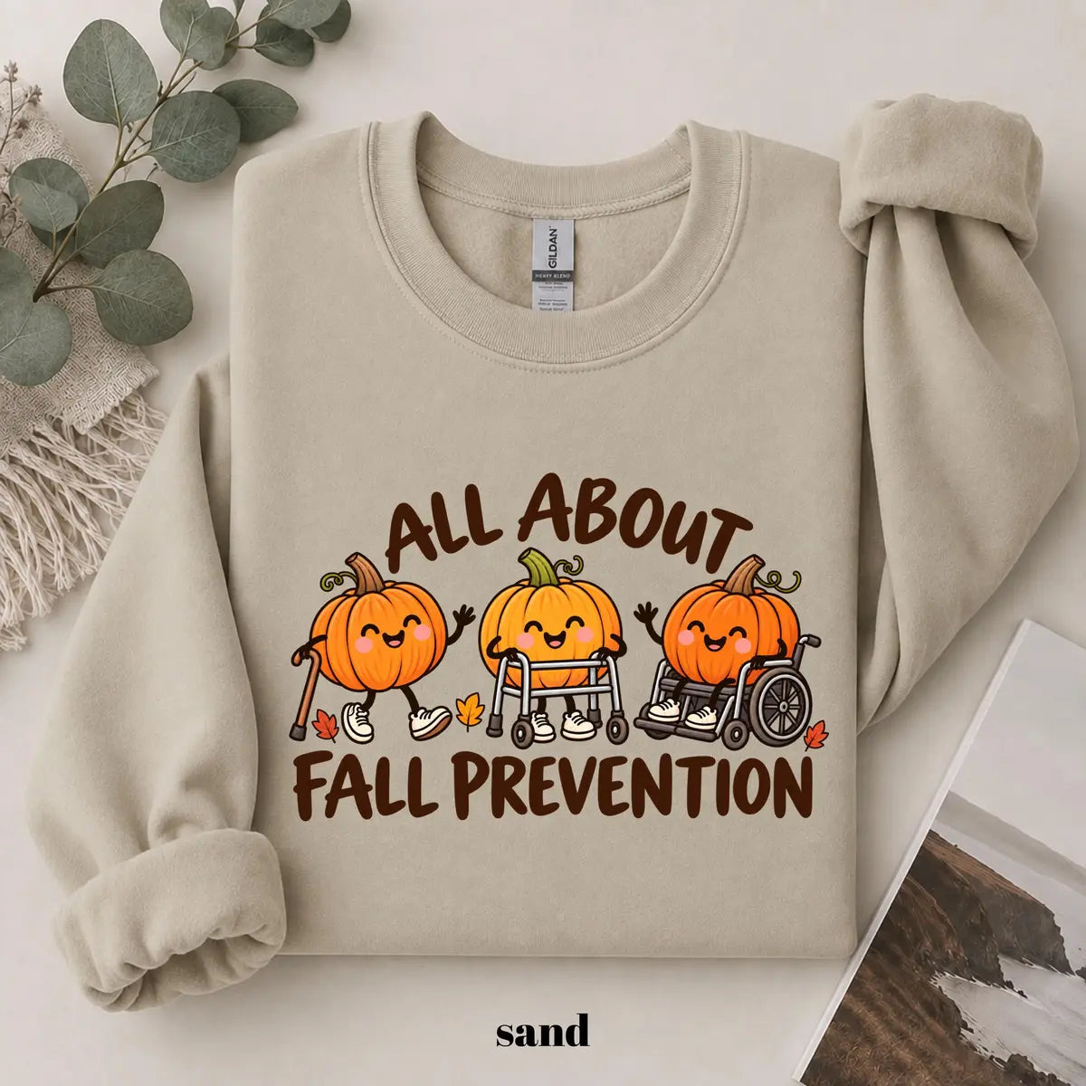 Sand crewneck sweatshirt with printed pumpkins with a walker and wheelchair above the words all about fall prevention, flat lay view.