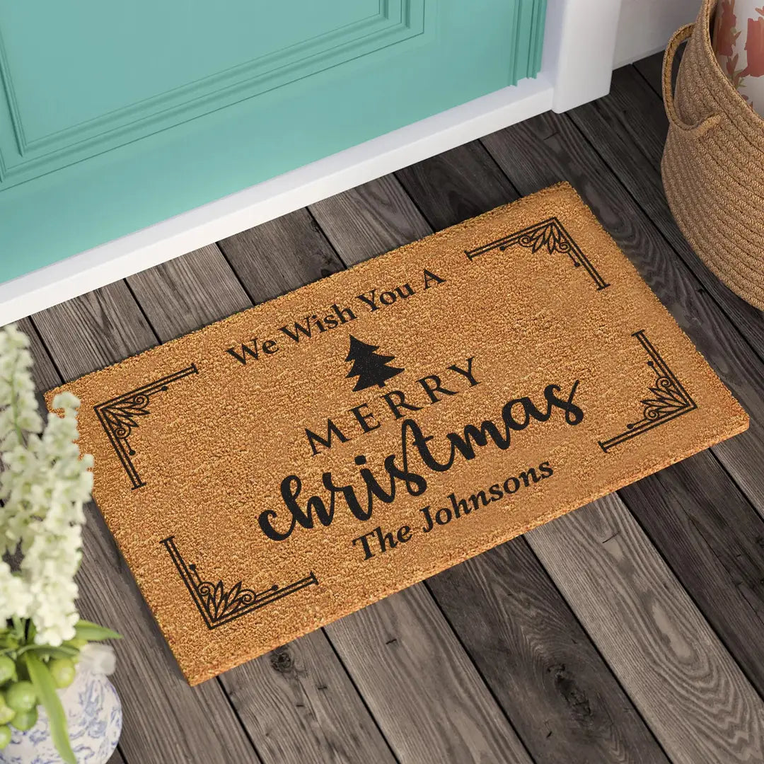 Coir doormat with a We Wish You a Merry Christmas message, pine tree, and family name on a wooden porch beside a teal door