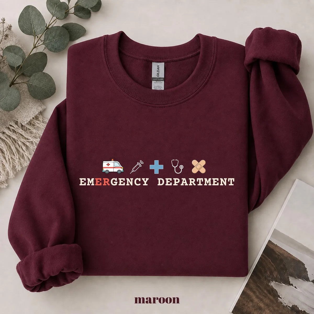 Maroon crewneck sweatshirt with printed an ambulance, syringe, cross, stethoscope and bandage above the words emergency department, flat lay view.