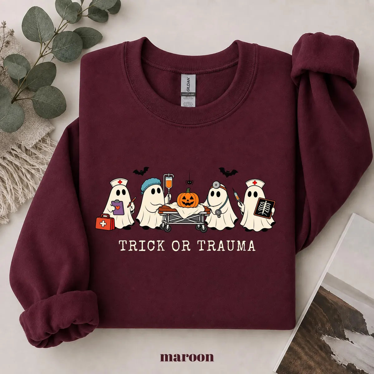 Maroon crewneck sweatshirt with printed nurse ghosts with a pumpkin and the words trick or trauma, flat lay view.