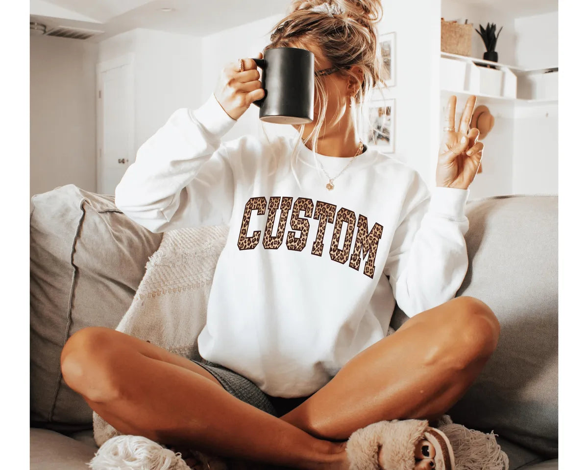 White crewneck sweatshirt with bold leopard print varsity lettering with your own custom name or text, worn by a model.