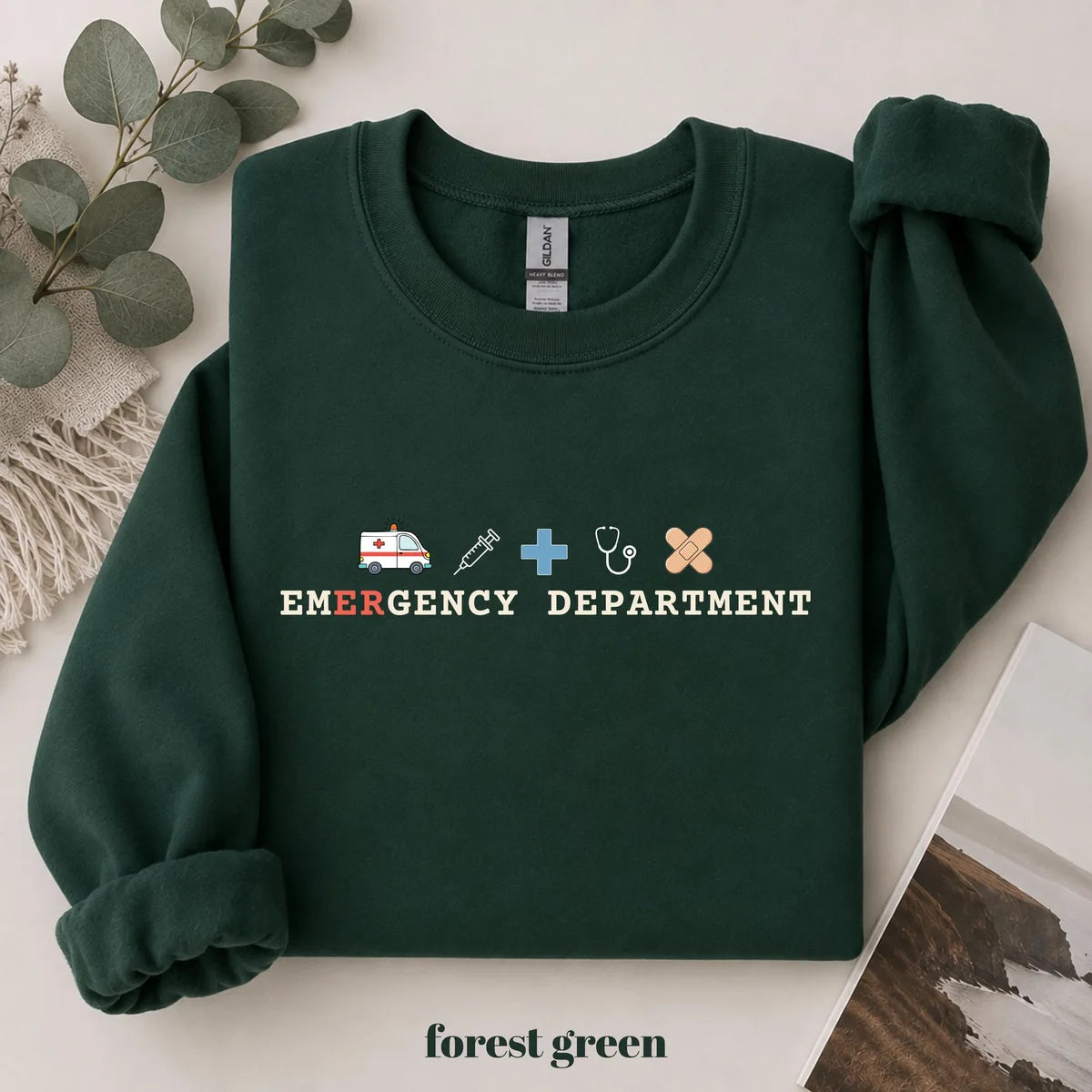 Forest Green crewneck sweatshirt with printed an ambulance, syringe, cross, stethoscope and bandage above the words emergency department, flat lay view.