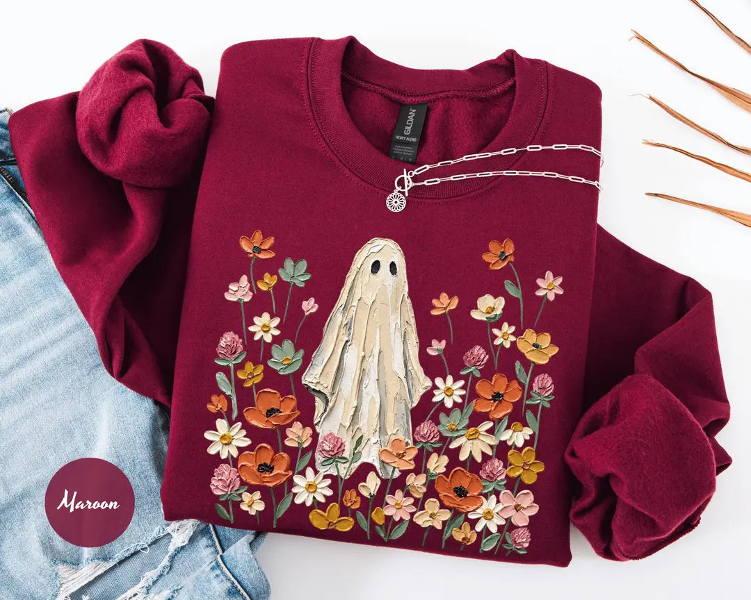 Maroon crewneck sweatshirt with printed a ghost standing in a field of autumn wildflowers, flat lay view.