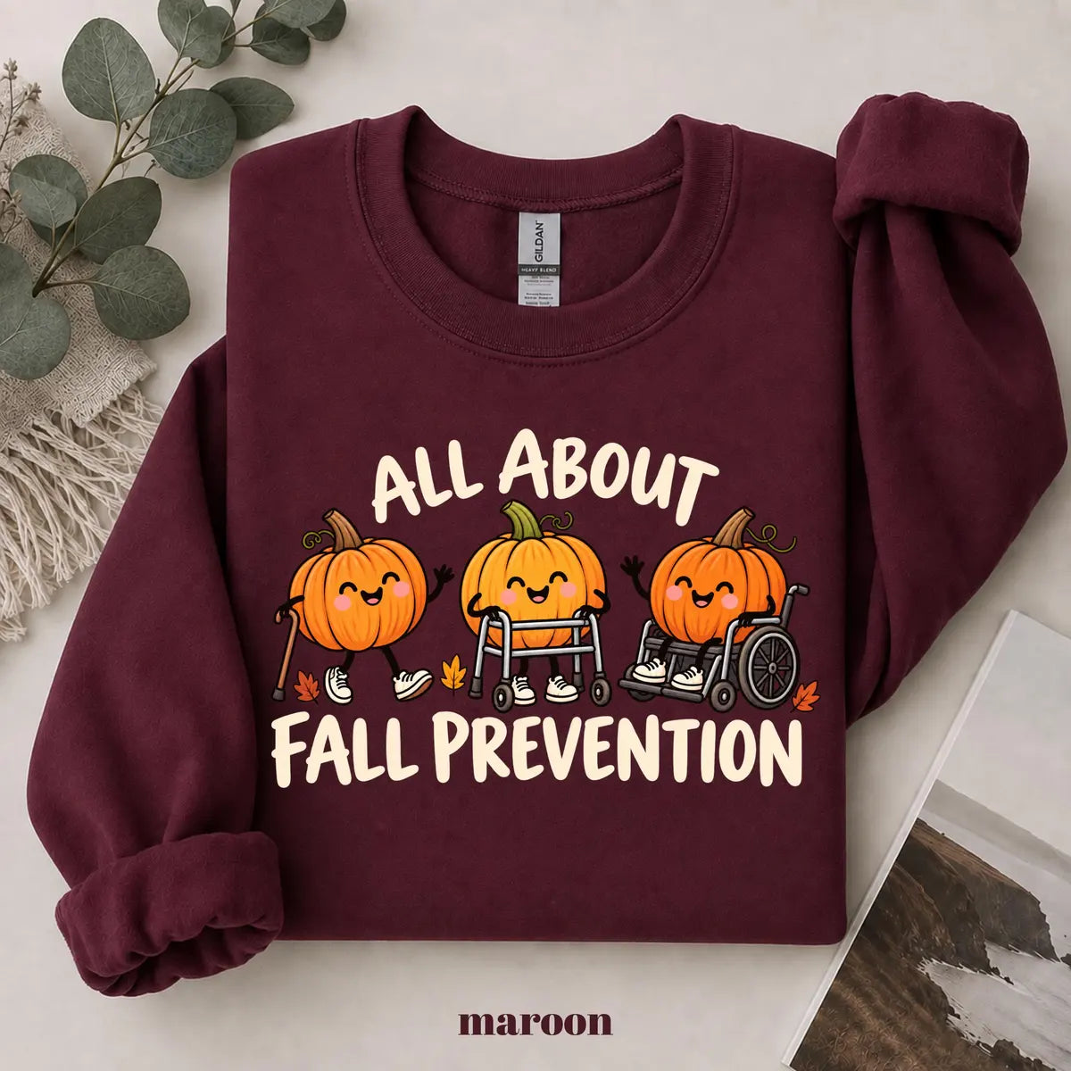 Maroon crewneck sweatshirt with printed pumpkins with a walker and wheelchair above the words all about fall prevention, flat lay view.