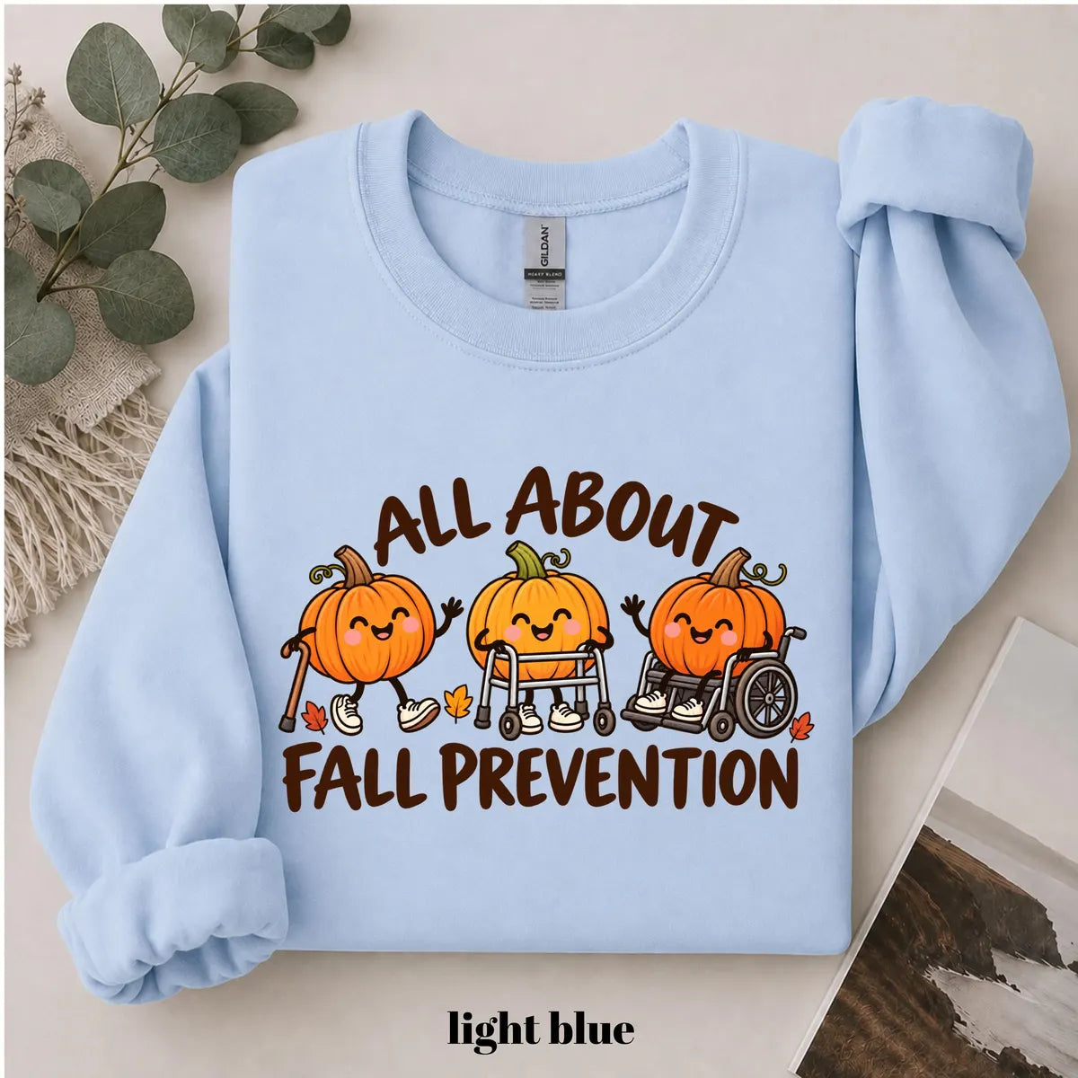 Light Blue crewneck sweatshirt with printed pumpkins with a walker and wheelchair above the words all about fall prevention, flat lay view.