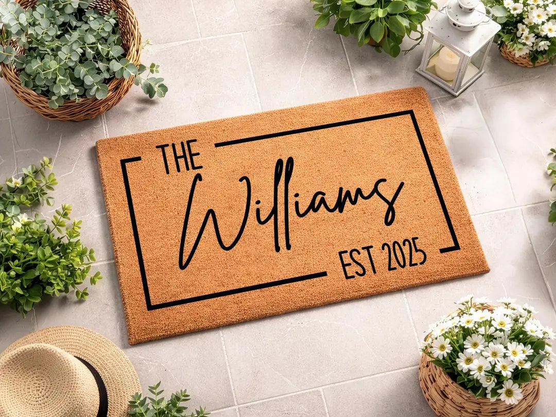 Coir doormat with a framed family name and established year on pale tile with a straw hat, flower baskets, and a lantern