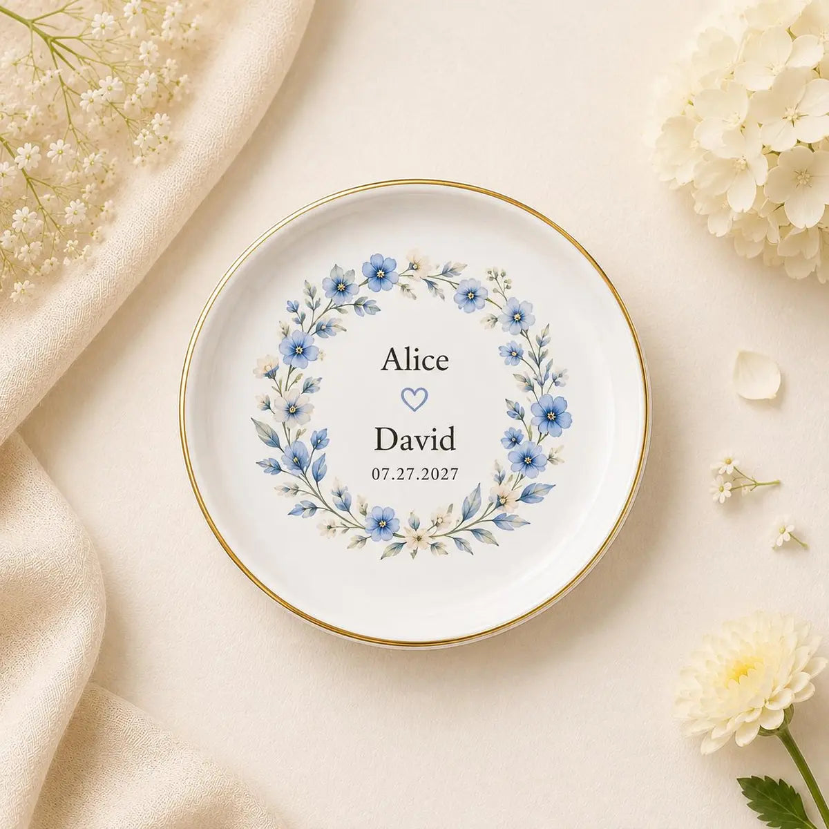 Round ceramic wedding ring dish with gold rim and a blue floral wreath around custom names and date