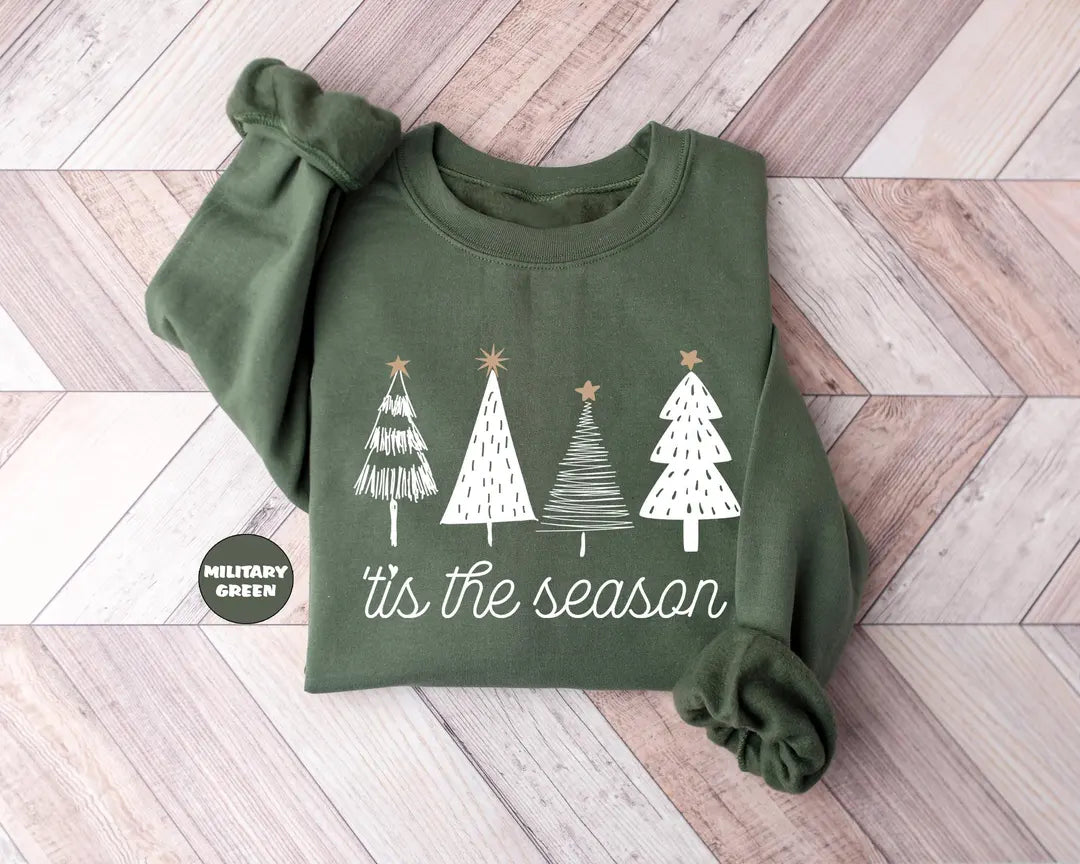 Military Green crewneck sweatshirt with four sketch-style Christmas trees above the script words tis the season, flat lay view.