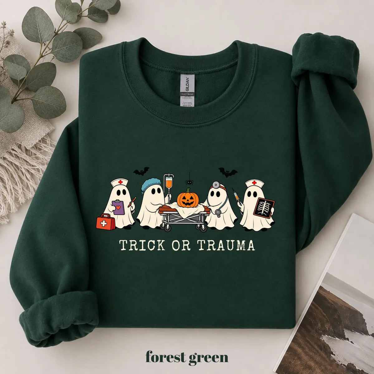 Forest Green crewneck sweatshirt with printed nurse ghosts with a pumpkin and the words trick or trauma, flat lay view.