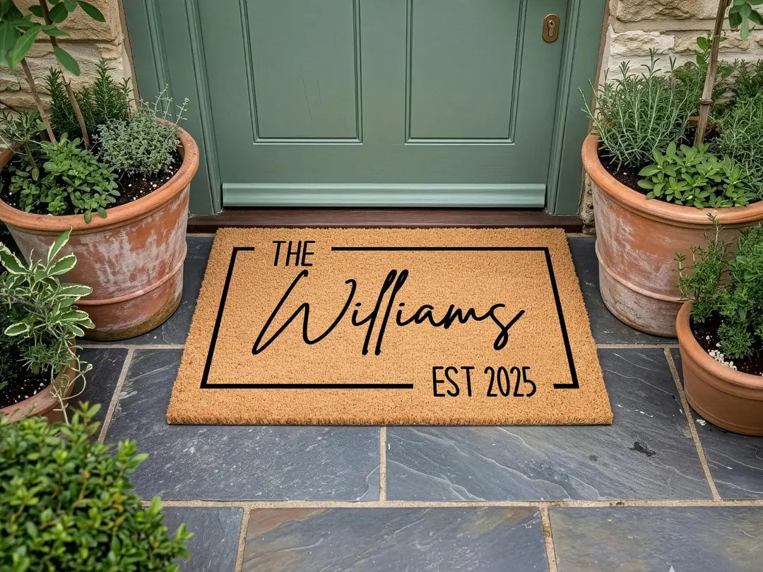 Coir doormat with a framed family name and established year at a sage green door with terracotta herb pots