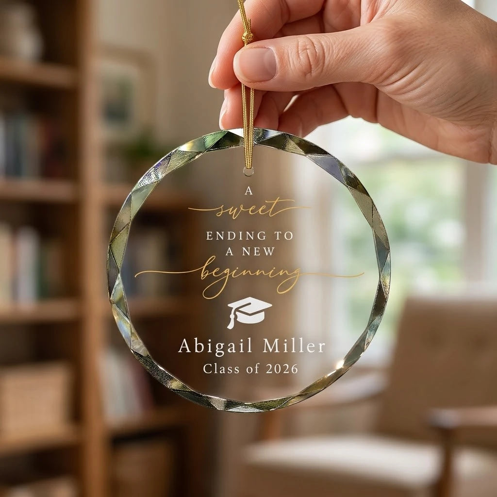 Hand holding a transparent faceted glass ornament with gold string loop showing printed graduation cap design and crisp white lettering in cozy indoor scene