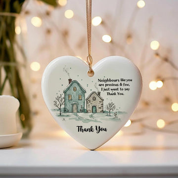 Heart-shaped ceramic ornament with a house illustration and text, hanging against a blurred background with lights.