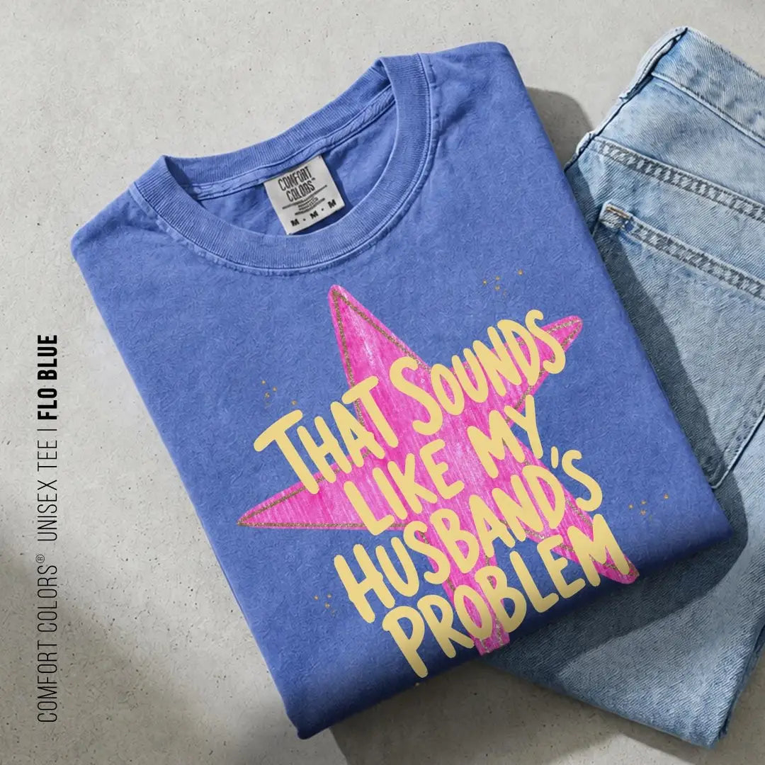 Folded Flo Blue Comfort Colors tee on blue jeans with yellow husband's problem lettering over a pink brush star