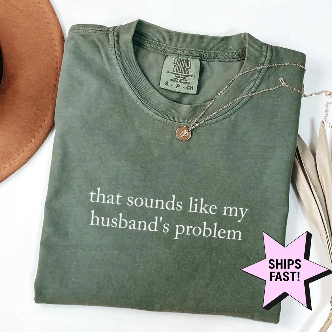 Folded Moss green Comfort Colors tee with the husband's problem quote in white serif, dried palm leaves nearby