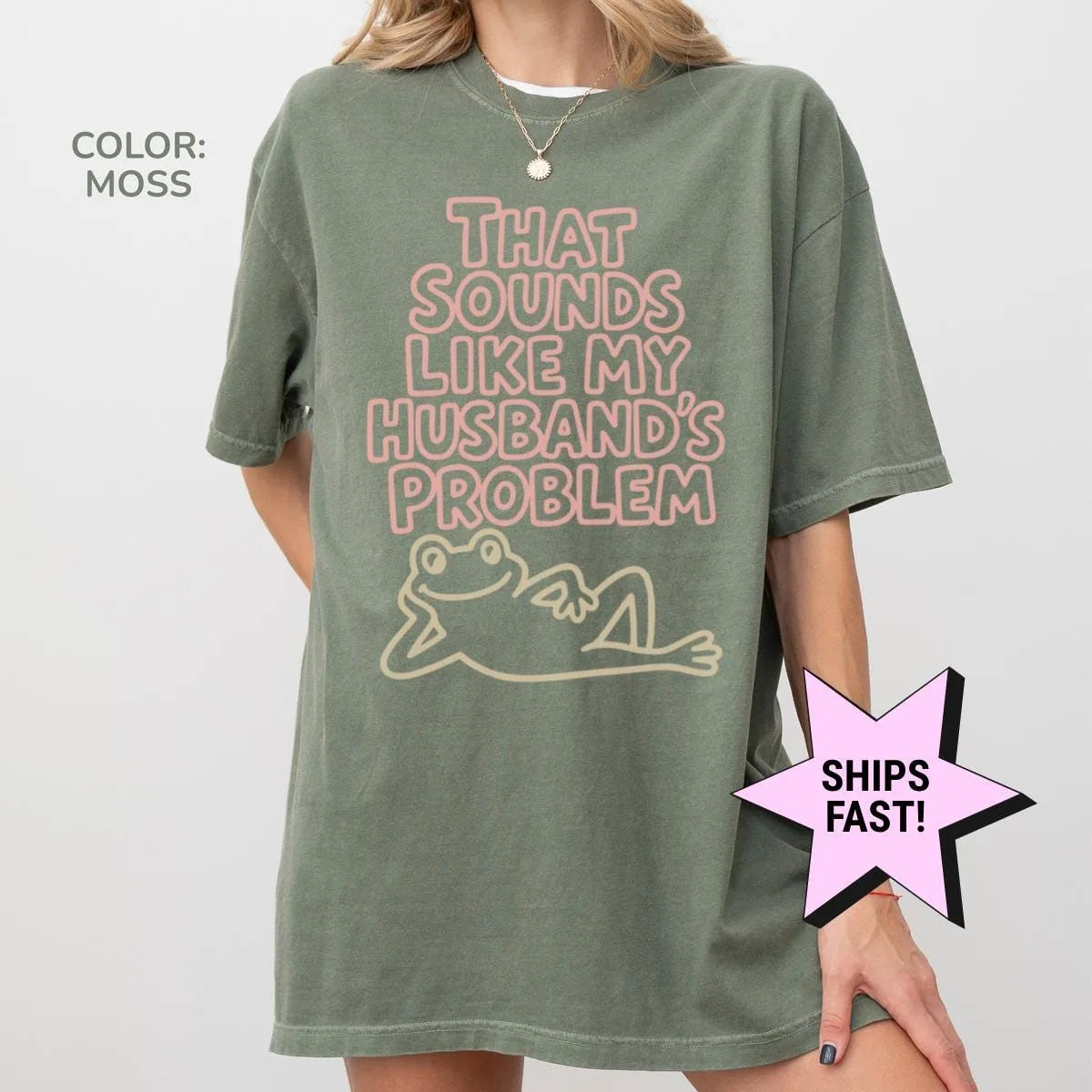 Woman in a Moss green Comfort Colors tee, hand on her knee, with pale pink husband's problem lettering and a cream frog
