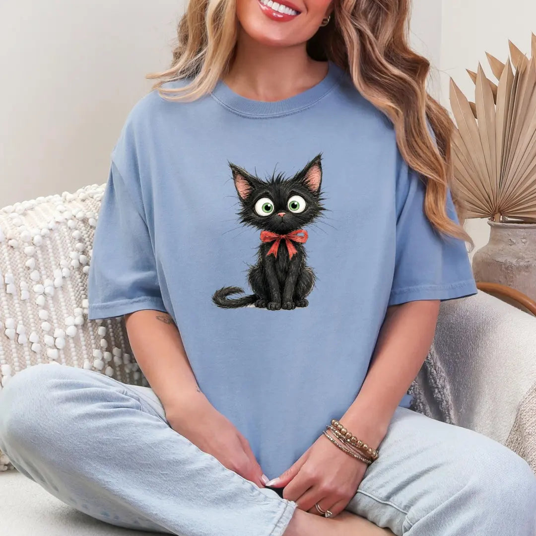 Woman in a Blue Jean Comfort Colors tee sitting cross-legged, a small scruffy black kitten with a red bow on the chest