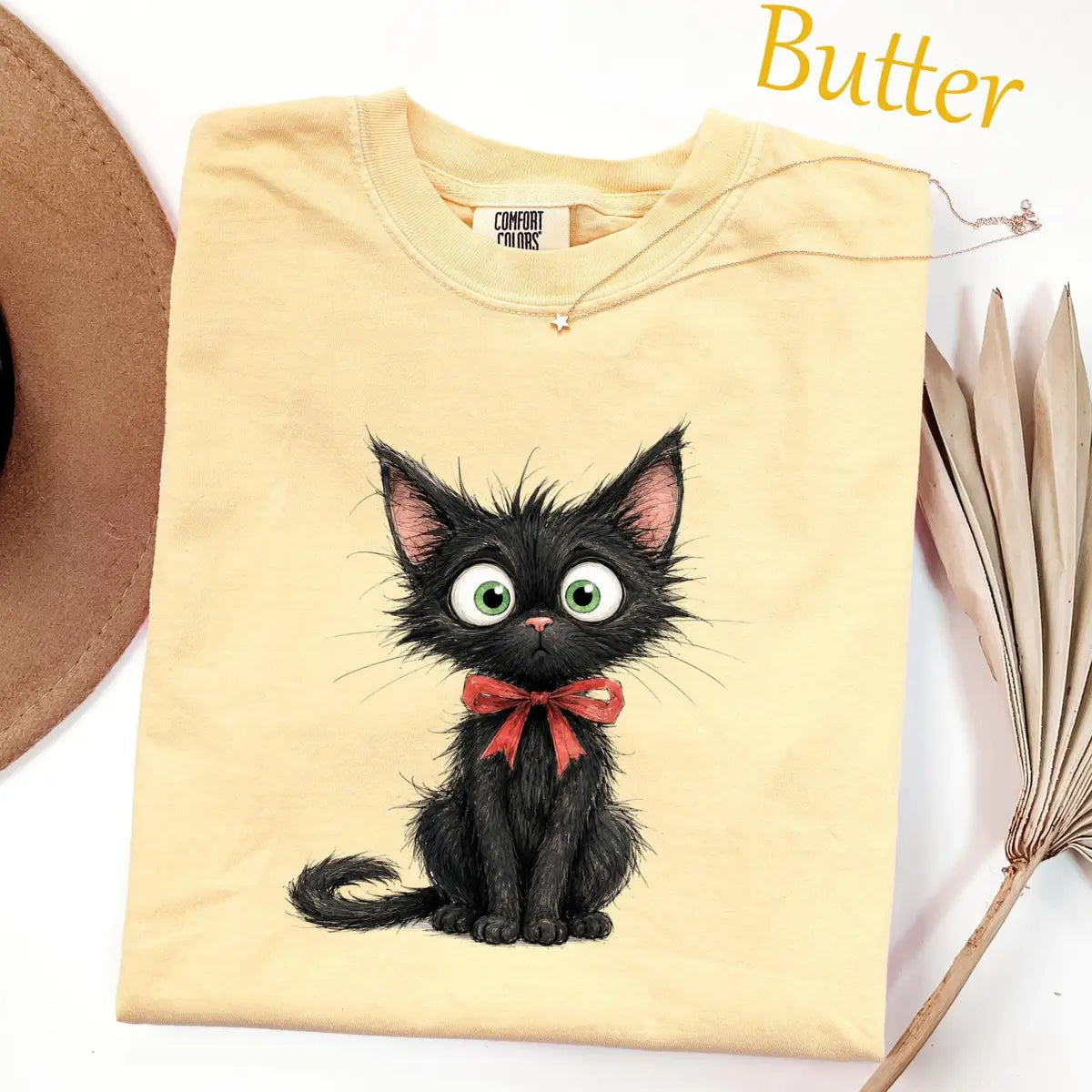 Folded Butter yellow Comfort Colors tee with a scruffy black kitten with big green eyes wearing a red bow, hat nearby
