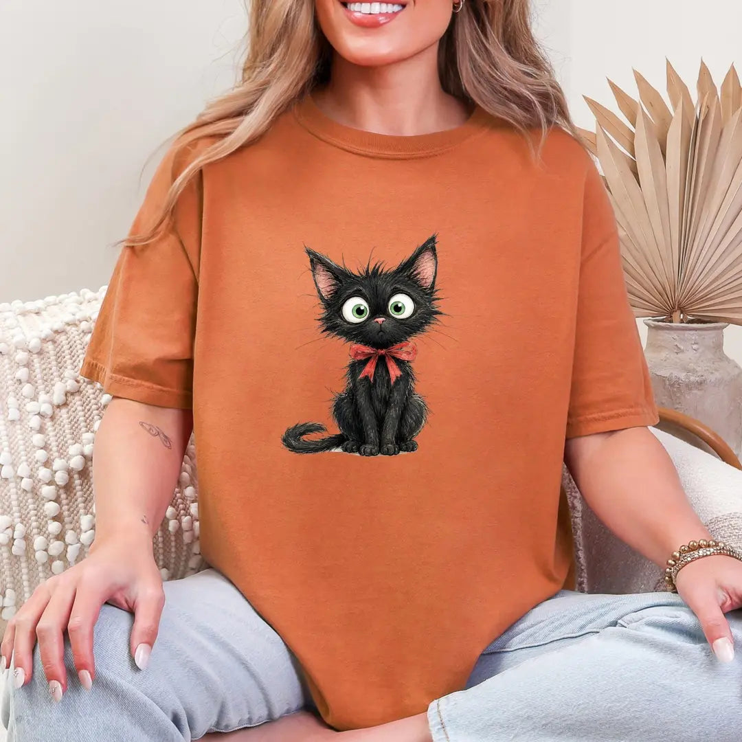 Woman in a Yam orange Comfort Colors tee sitting on a couch, a small black kitten with green eyes and a red bow