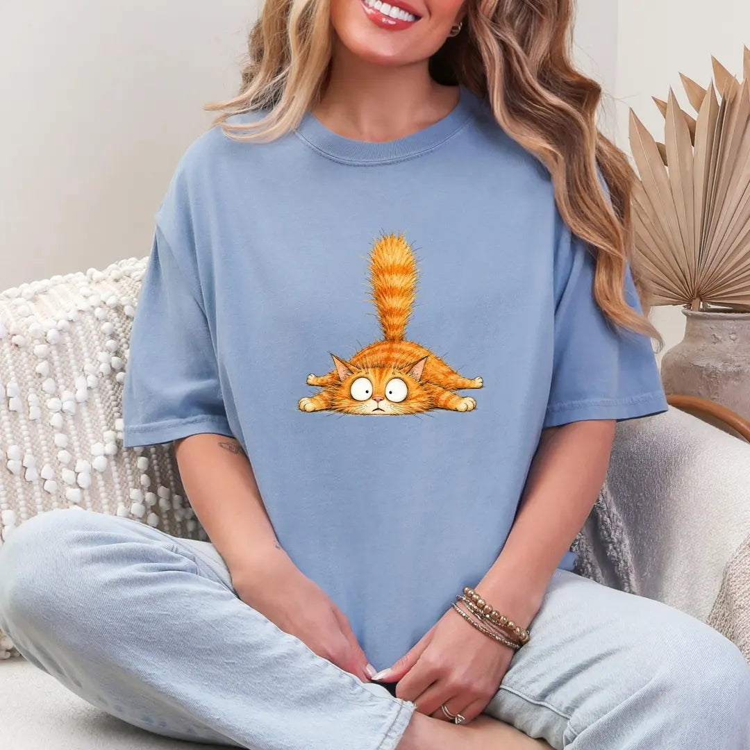 Woman in a Blue Jean Comfort Colors tee sitting cross-legged, a small orange tabby with googly eyes on the chest