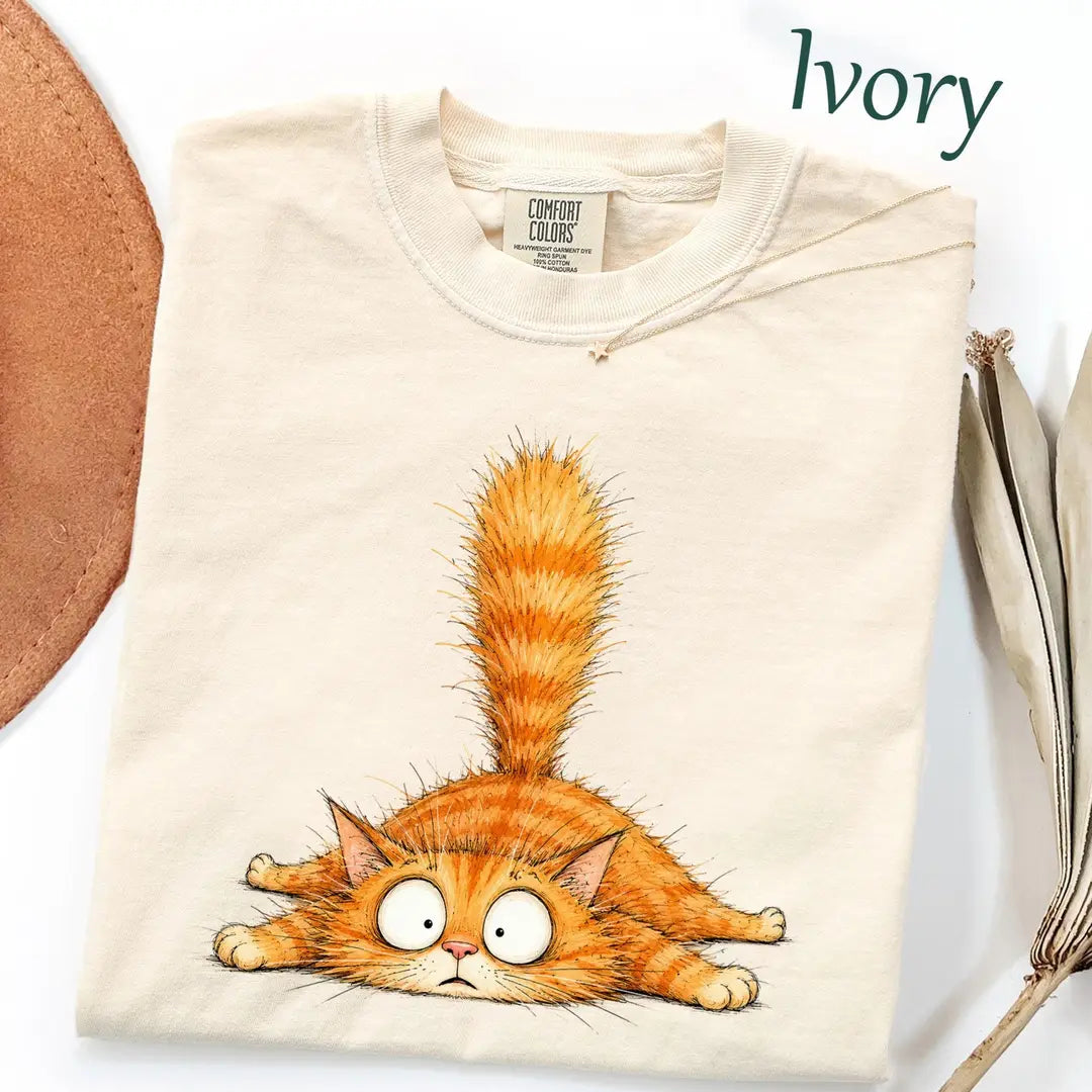 Folded Ivory Comfort Colors tee with a fluffy orange tabby lying flat, wide googly eyes and its tail straight up, hat nearby