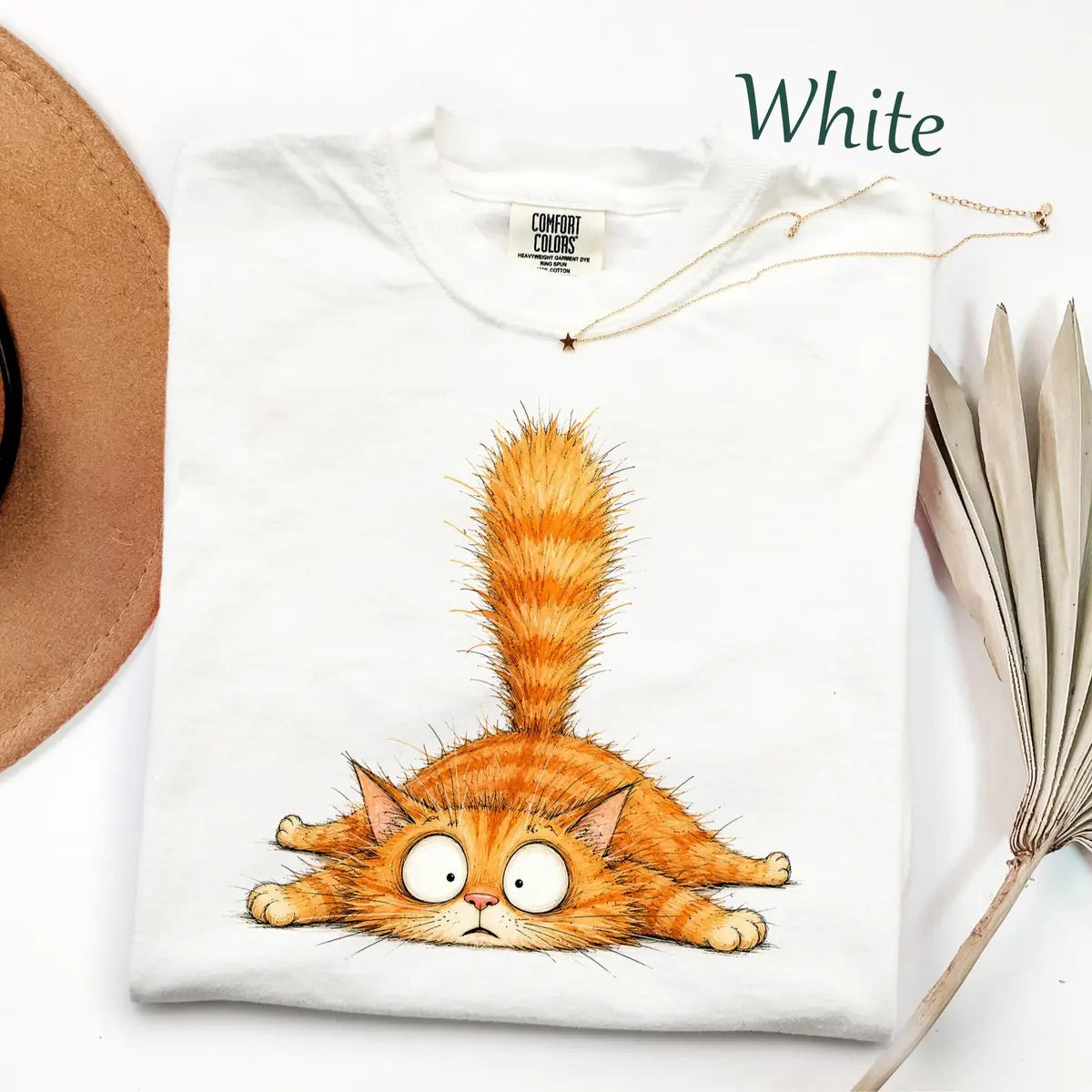 Folded White Comfort Colors tee with a fluffy orange tabby lying flat, wide eyes and its tail up, necklace on the collar